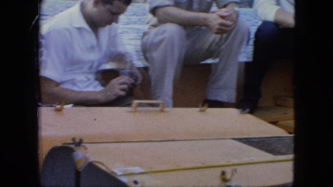 Adults sit along a wooden boat with water in the background, showing casual travel clothing and deck details in a 1960s home-movie frame.