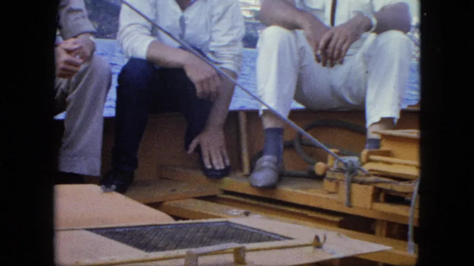 Close 8mm frame of passengers' legs and footwear on a wooden boat, with rope, rod, and deck hardware visible during a sea crossing.