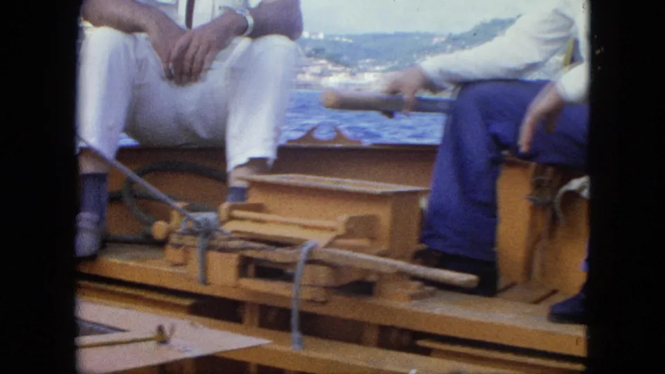 Seated adults on a small wooden boat near water and a distant shoreline, with ropes and deck fittings visible in the foreground.