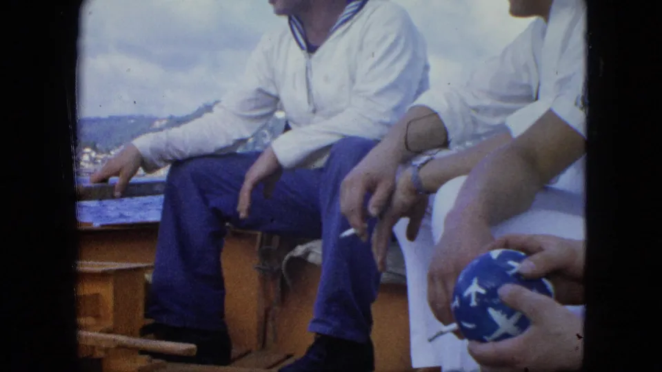 1964 8mm frame of seated adults on a wooden boat, including a cigarette in hand, a blue patterned ball, open water, and distant shore.