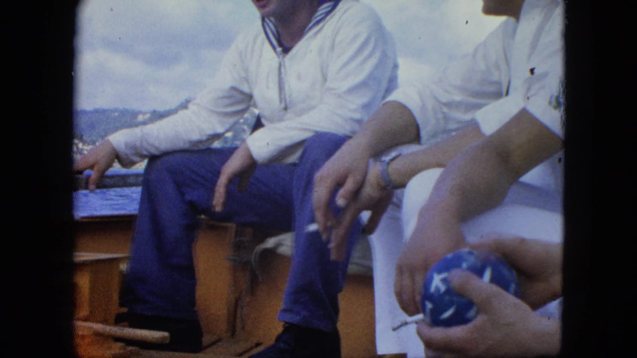 Two men in white tops sit on a ship deck, with a hand holding a blue patterned ball in the foreground.