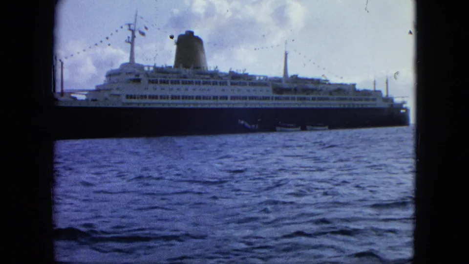 Handheld 8mm shot of a large passenger liner broadside across rippling water, with upper decks and rigged lights visible.