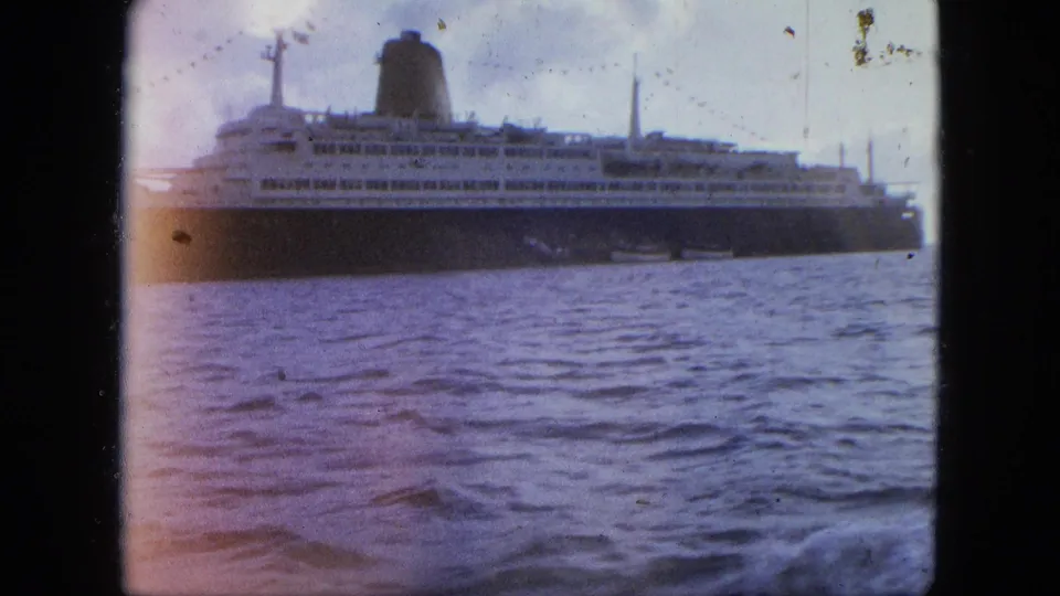 8mm frame of the ocean liner at sea with visible water, cloudy sky, and a warm light leak across the left side of the frame.