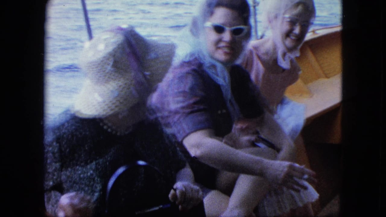 Three women seated in a small boat, with water behind them and one woman leaning forward in a straw hat.