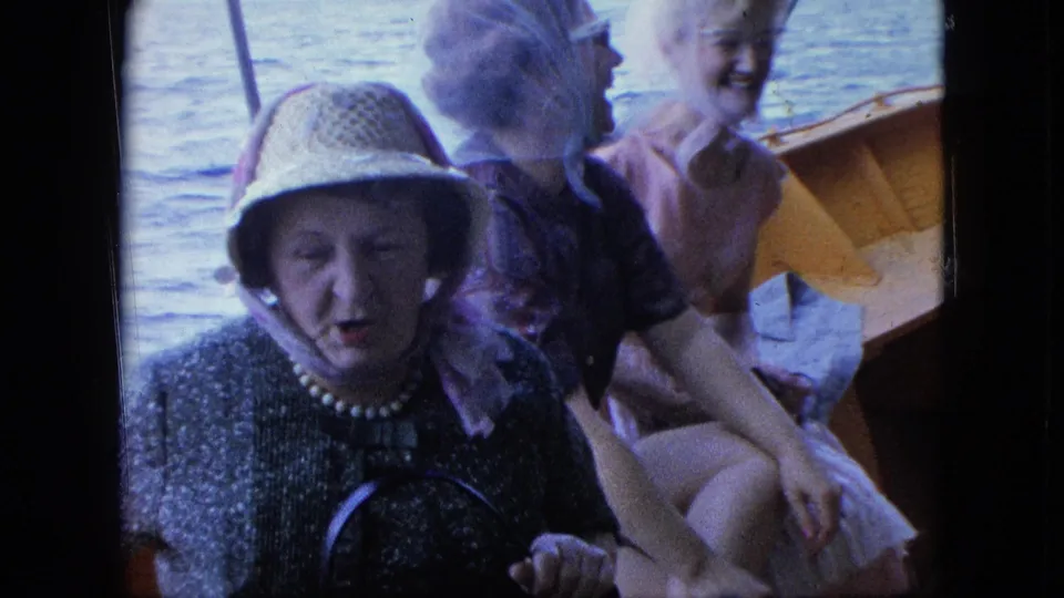 Home-movie frame of an older woman in a straw hat and pearl necklace seated before two smiling women on a boat.