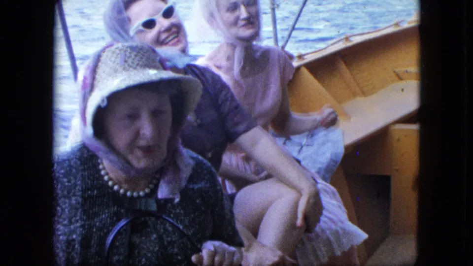 Close 8mm frame of three women seated on a small boat, with sunglasses, a straw hat, and pale dress visible against blue water.