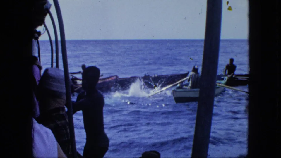 Handheld boat view looking across open water, with dark foreground figures and distant small boats near a splash or wake.