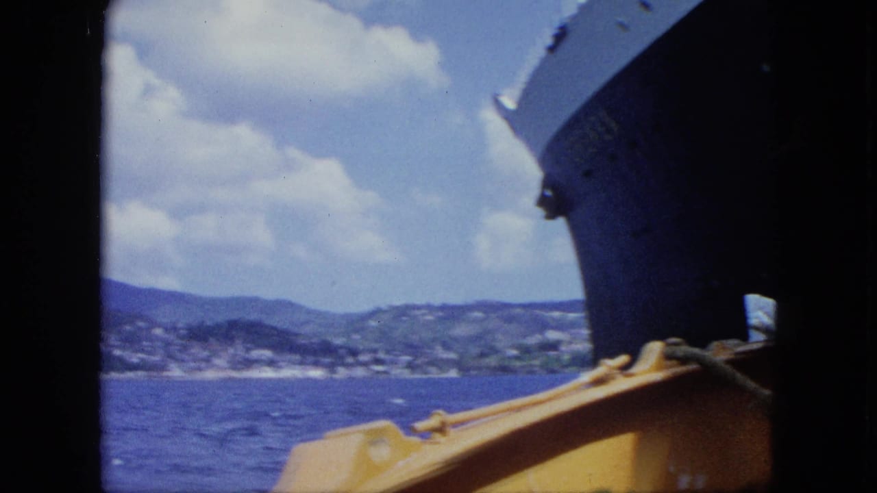 View from a small boat near the dark bow of a large liner, with blue sea, distant tropical hills, clouds, and yellow boat structure in frame.
