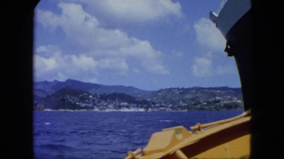 1964 home-movie view across blue water toward a hilly coastline, with the edge of the ship and yellow small-boat structure framing the scene.