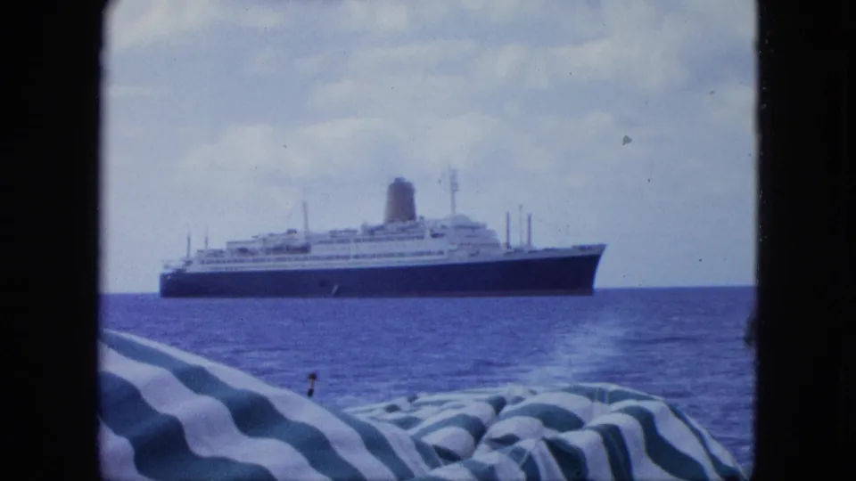Distant view of a large ocean liner offshore, filmed from a small boat with striped fabric in the foreground and open sea around it.