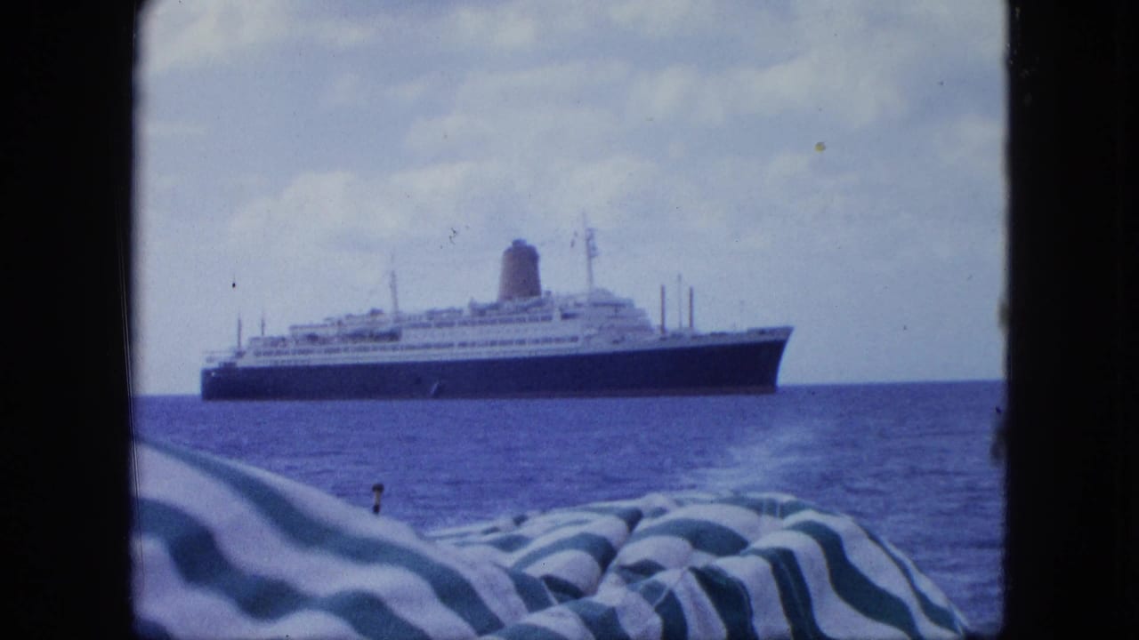 Dark-hulled passenger liner seen across calm blue water from a nearby vessel, with striped deck fabric at the lower edge.
