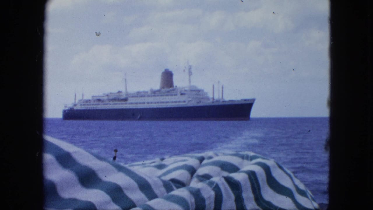 Large ocean liner with a dark hull and pale decks viewed from a boat, framed by blue sea, cloudy sky, and striped fabric.