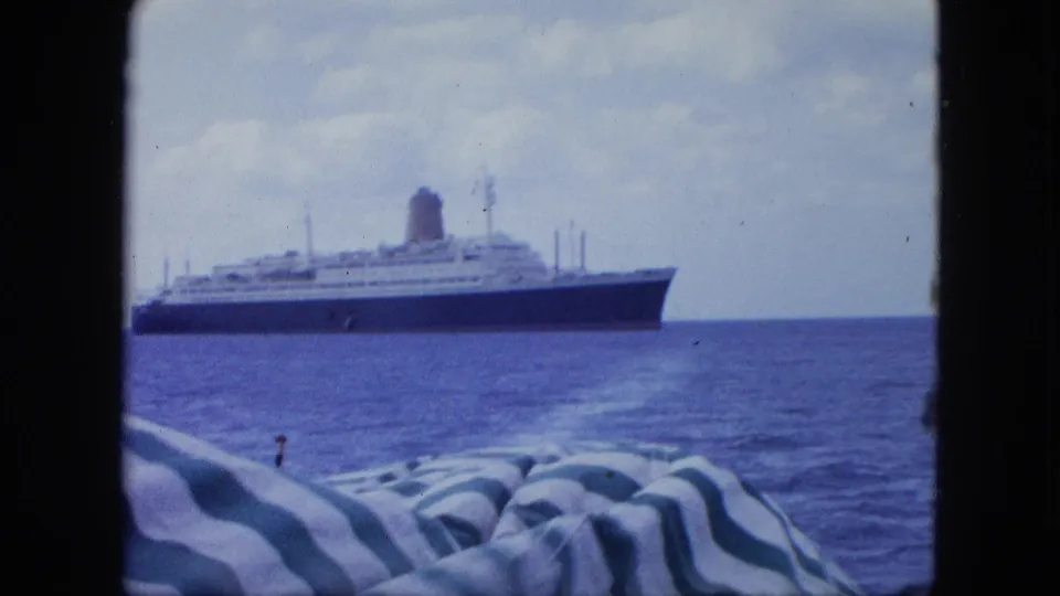 Passenger ship passing across calm water, with striped fabric and a small deck fitting visible from the filming boat.