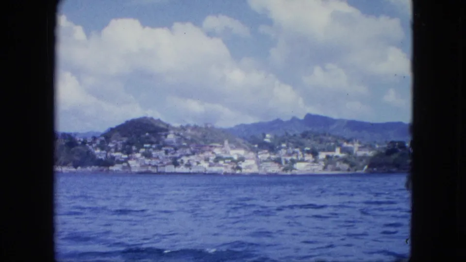 Distant coastal town built along a hillside across blue water, seen from a boat under a partly cloudy sky.