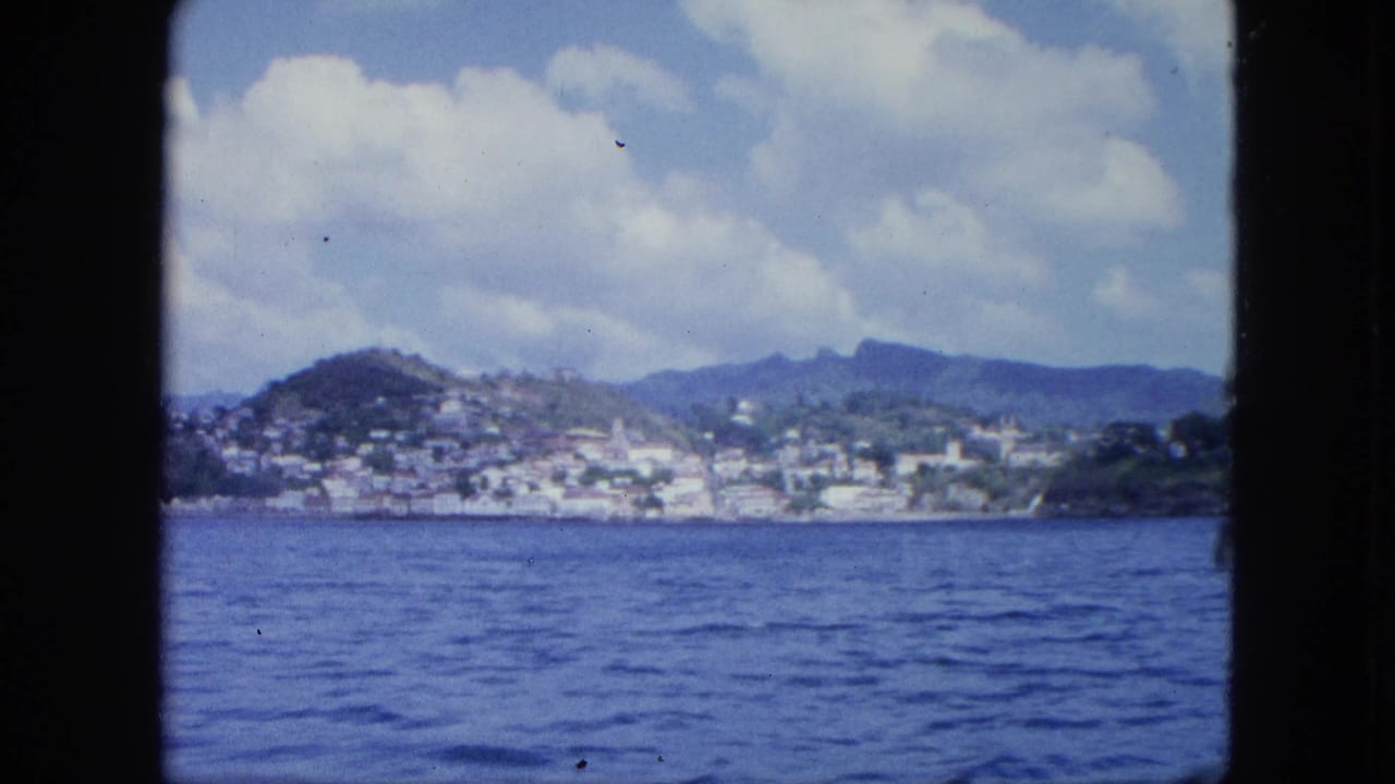 Soft 8mm frame of blue water, a distant coastal settlement, green hills, and mountain ridges seen from a vessel.
