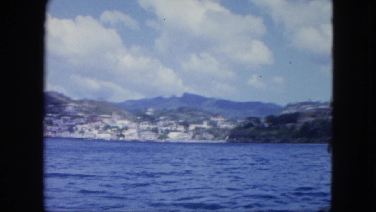 Shipboard-looking 8mm view across blue sea toward a hillside settlement and mountains beneath a cloudy sky.
