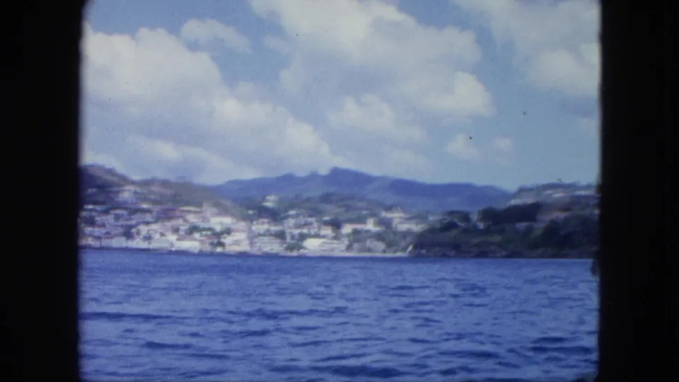 Shipboard-looking 8mm view across blue sea toward a hillside settlement and mountains beneath a cloudy sky.