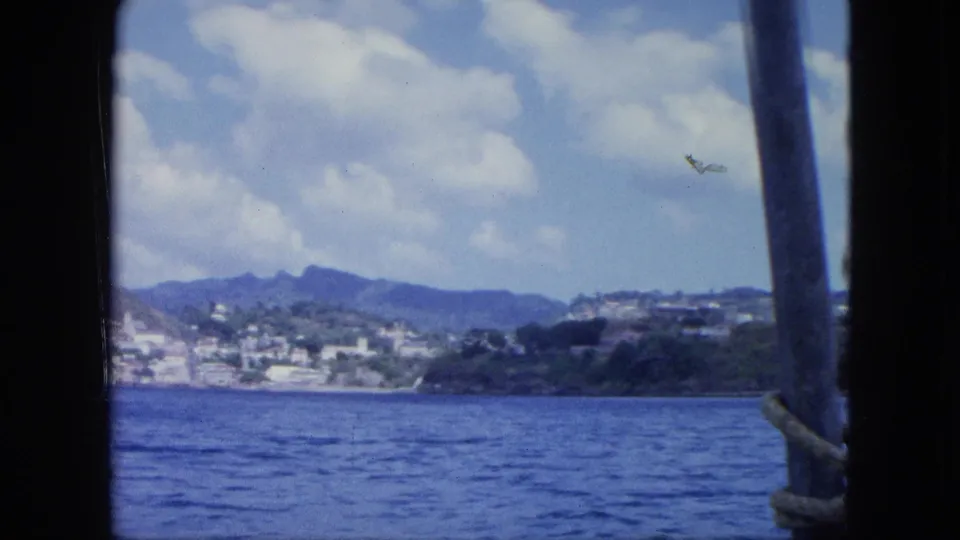 8mm travel frame showing blue water, a distant coastal slope, mountain ridges, and part of a ship structure in the foreground.