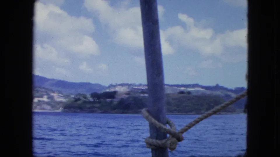 Foreground ship post and rope frame a distant island coastline with blue water, low hills, and scattered clouds.