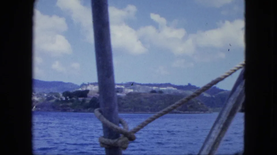 1964 8mm home-movie view from a boat, with rope and a pole framing blue water and a distant green Caribbean coastline.