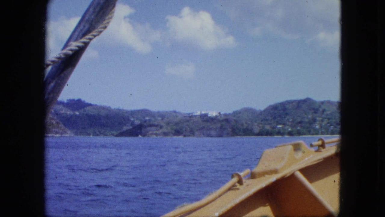 1964 8mm frame showing open water, distant hills, sky, and the yellow edge of a boat near a Caribbean shoreline.
