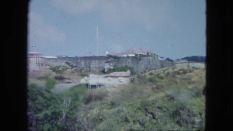 Cataloged 1964 home-movie frame showing a fortified hillside site, small buildings, scrub vegetation, and sky.