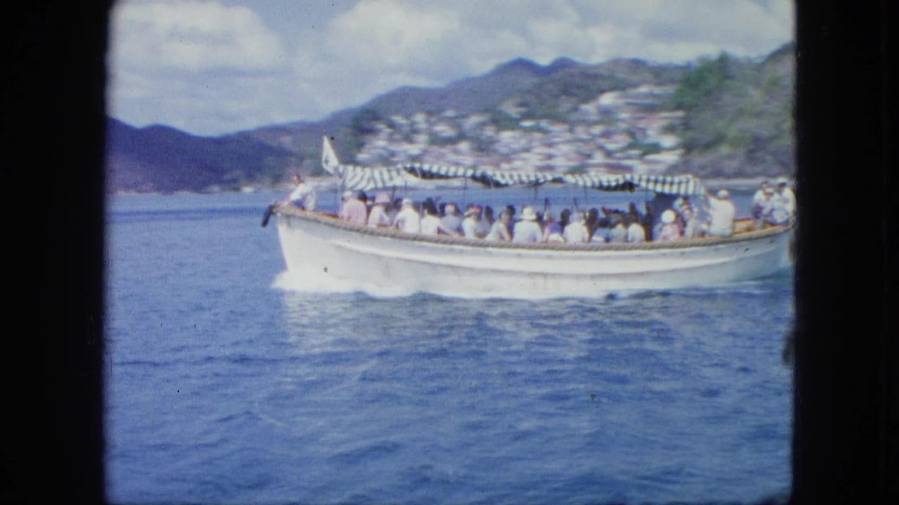 A white tour boat carrying seated passengers under a striped canopy passes across blue water near a hilly coastal town.