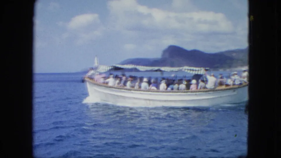 Side view of a crowded white sightseeing boat under a striped canopy, filmed from another vessel or shoreline across blue water.