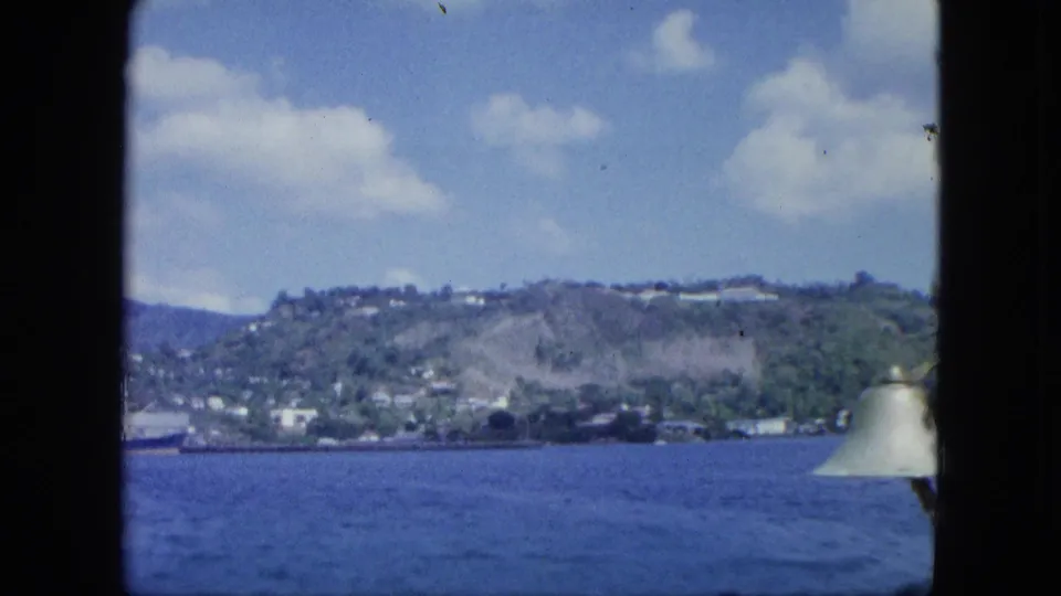 A hilly coastal settlement and green slopes are seen across blue water, with a boat fixture entering the right edge of the frame.