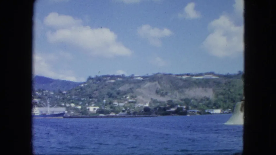 1960s 8mm home-movie view from a boat toward blue harbor water, hillside buildings, a docked ship, and scattered clouds.