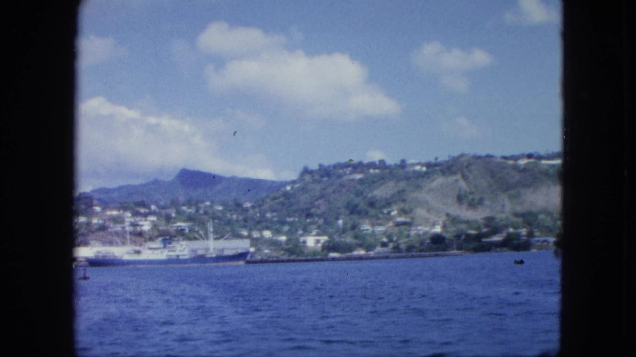 Hillside shoreline and docked ship across blue water under a bright cloudy sky.