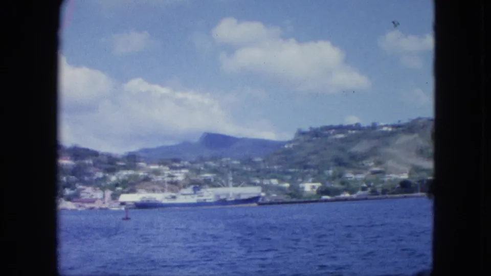 Vintage boat-shot harbor scene with rippled water, a docked cargo ship, hillside homes, distant landforms, and soft 8mm color.