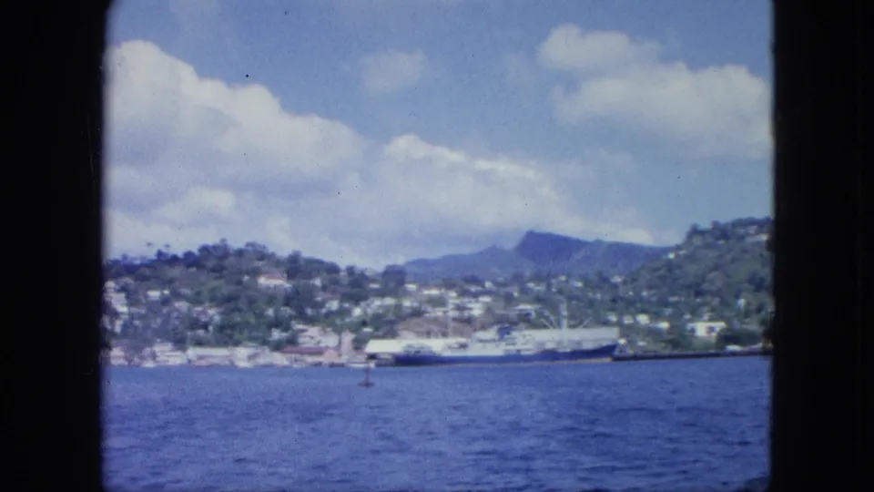1960s home-movie frame of a broad harbor view from a boat, with hillside buildings, a port vessel, distant peak, and cloudy sky.