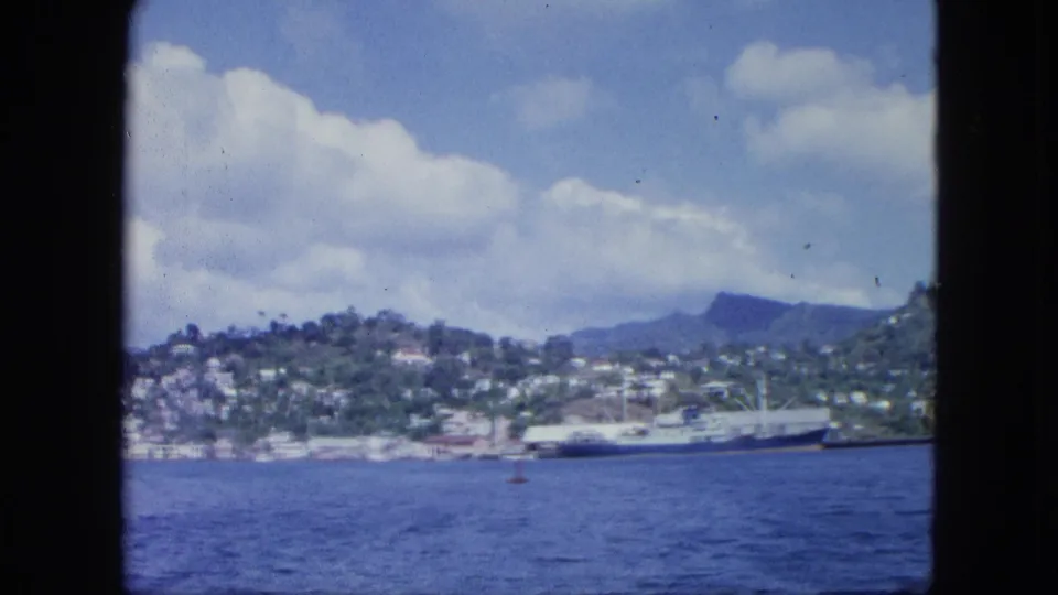 1964 8mm view from a vessel window of a hillside coastal town, blue harbor water, clouds, and a docked cargo ship.