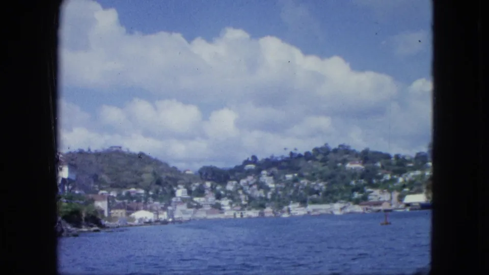 1964 8mm coastal view showing blue water, a low shoreline town, green hills, and a small marker as the vessel moves along the bay.