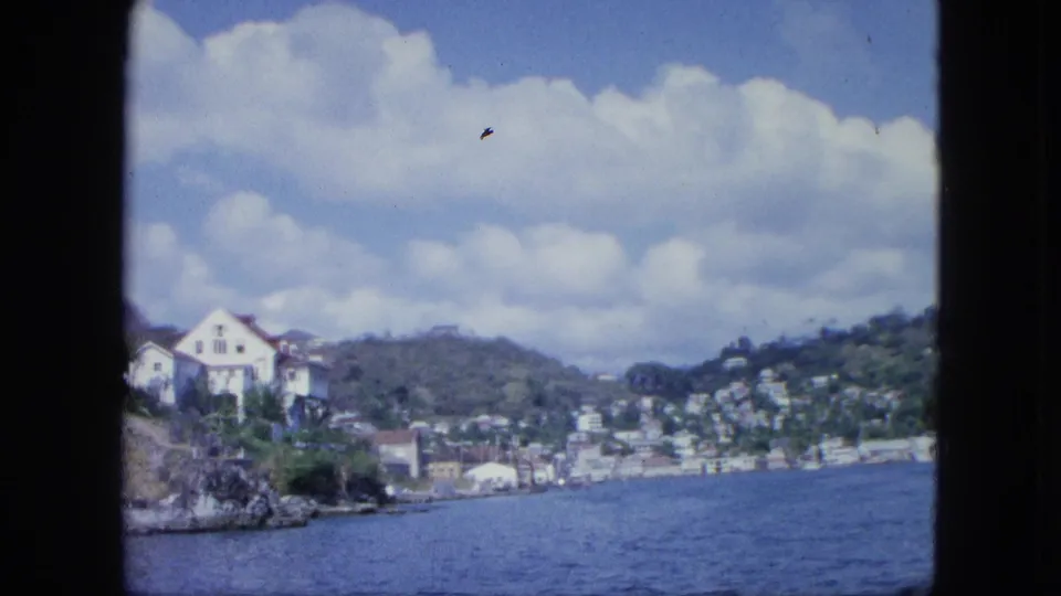 1964 8mm frame with a white house on a rocky waterfront point, blue water, hillside buildings, and clouds viewed from a moving vessel.