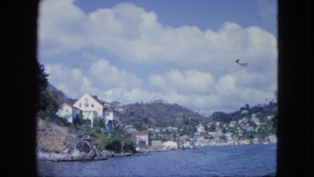 1960s 8mm boat view of a white hillside house, coastal water, scattered buildings, green slopes, and large clouds.