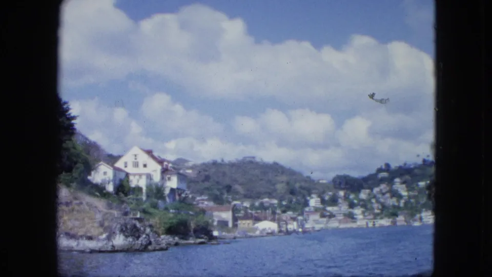 1960s 8mm boat view of a white hillside house, coastal water, scattered buildings, green slopes, and large clouds.
