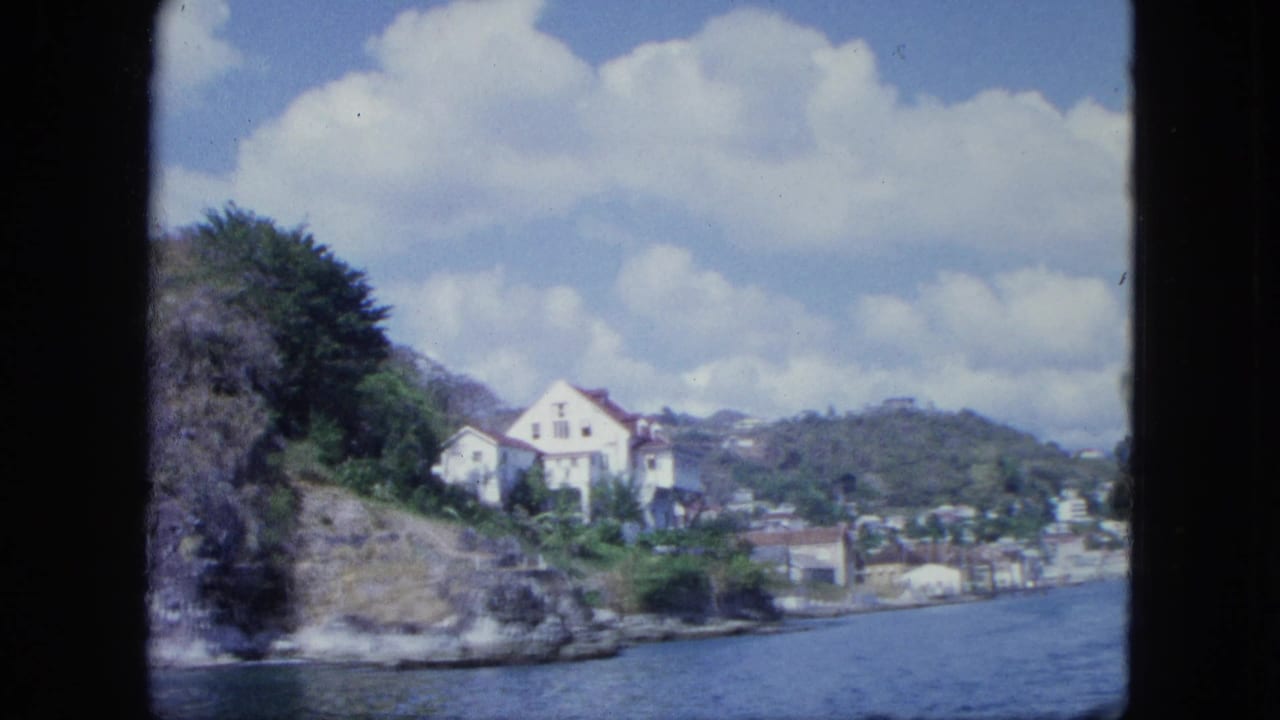 Boat-shot 8mm view of a white house on a rocky green shoreline, with harbor water, distant settlement, and clouds.