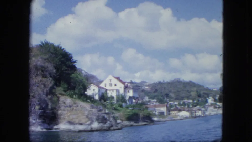 Boat-shot 8mm view of a white house on a rocky green shoreline, with harbor water, distant settlement, and clouds.