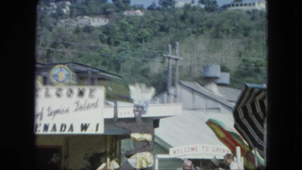 Close boat or street view of a Grenada, W.I. welcome sign, flags, low buildings, hillside greenery, and people near the lower edge.