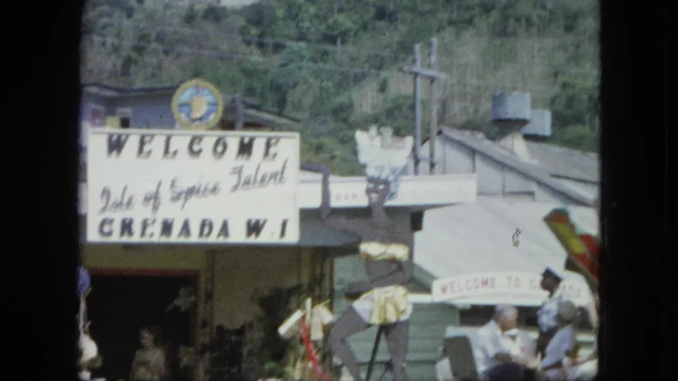 1960s 8mm view of a Grenada, W.I. welcome sign, decorative emblem, flags, waterfront buildings, and visible people near the entrance.