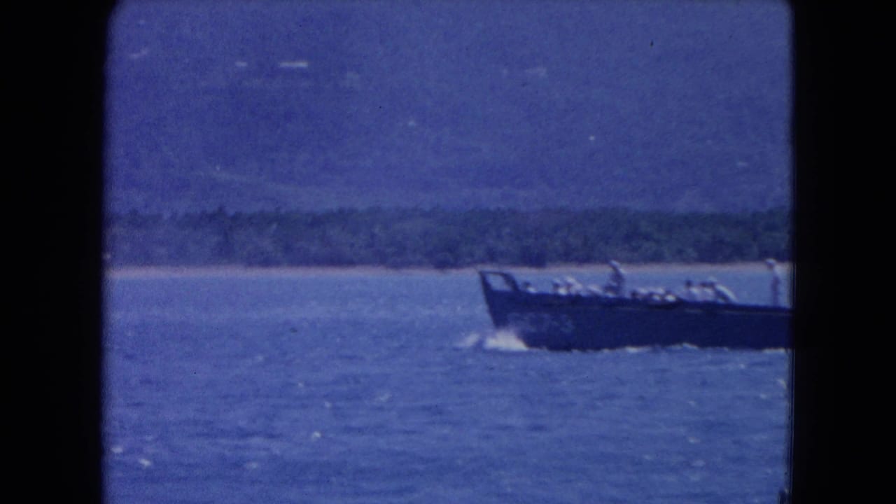 Dark landing craft on blue water with distant shoreline and mountains.