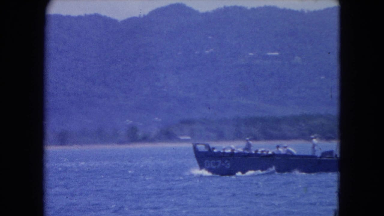 Landing craft with people aboard passes in front of a hazy coastline.