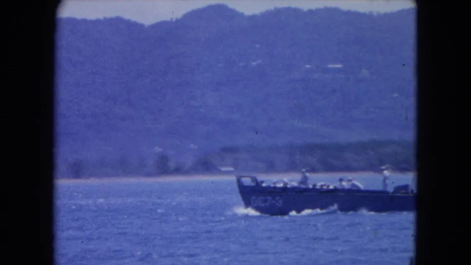 A military-style boat with several visible passengers moves offshore near Montego Bay, Jamaica, in soft 8mm color.