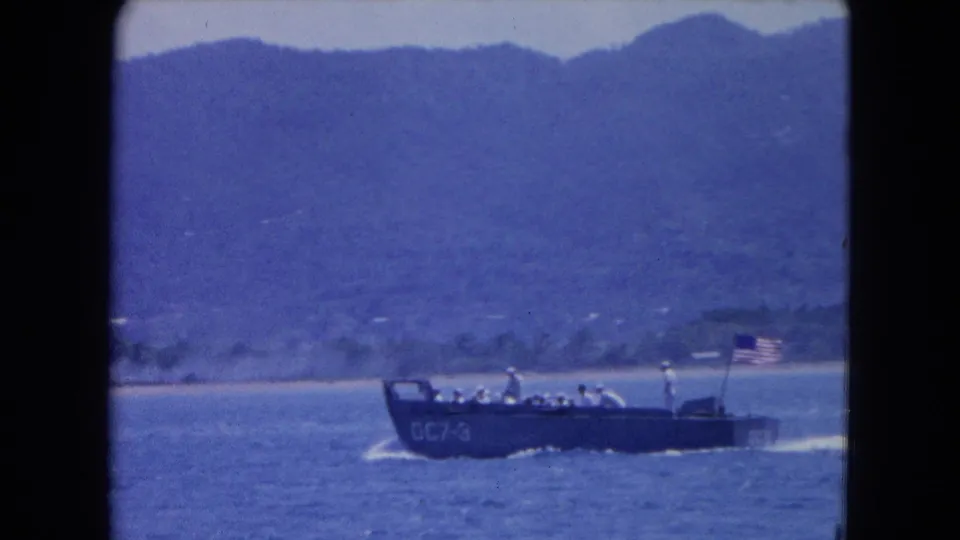The landing craft marked GC7-3 carries people and an American flag across the water near Montego Bay in 1964 footage.