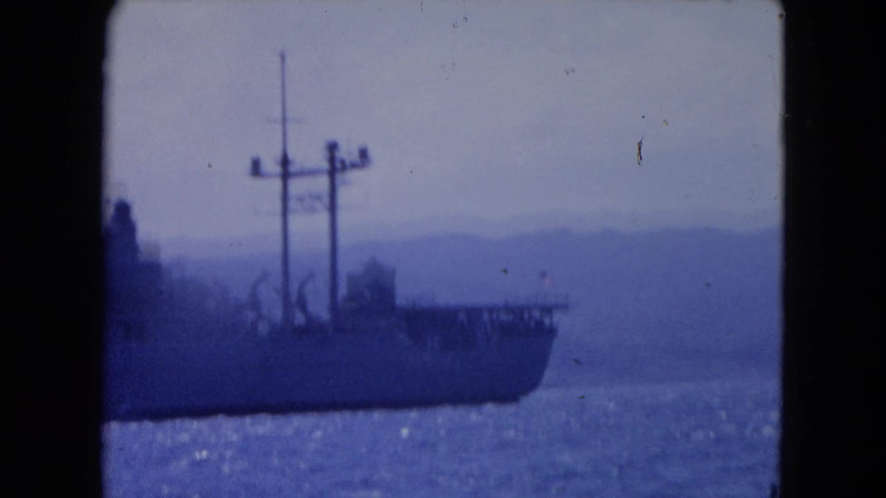 Large ship hull with cranes and antenna masts on blue water with distant hills.