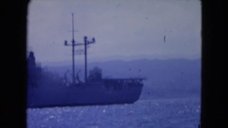 1964 Montego Bay 8mm frame showing a large ship partly in view, with deck cranes, masts, open water, and hazy coastal hills.