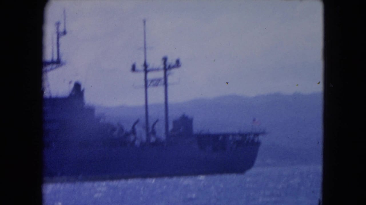 Ship stern area with masts and cranes silhouetted against sea and hills.