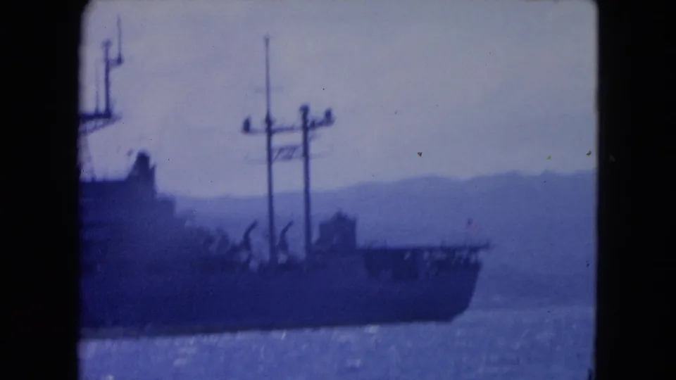 1964 Montego Bay home-movie frame with the aft section of a large ship, visible masts, cranes, blue sea, and distant shoreline.